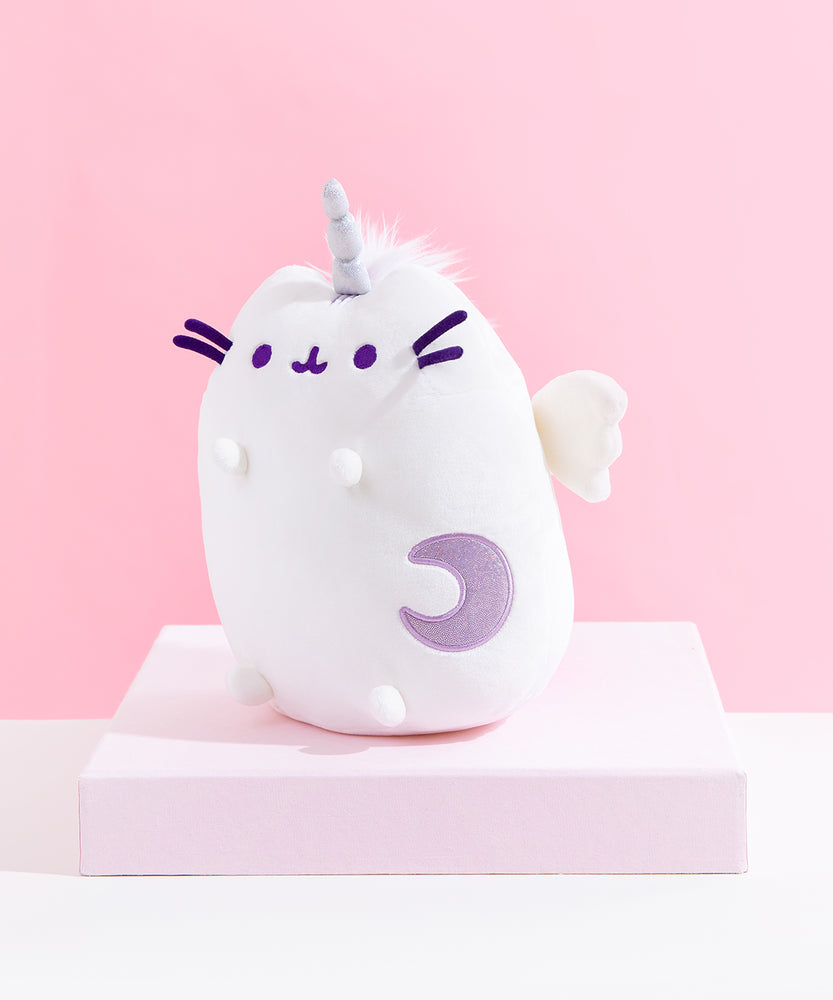 Quarter view of the Super Pusheenicorn Plush facing the left on top of a white square pedestal, in front of a pink background. The sparkly purple crescent moon on Pusheen’s front and the plush wing is more visible from this angle, though the multicolor mane is only visible directly behind Super Pusheenicorn’s horn.