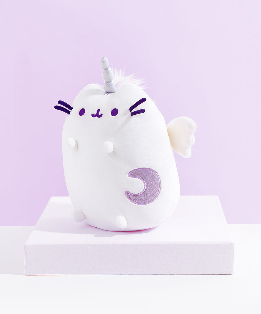Quarter view of the Super Pusheenicorn Plush facing the left on top of a white square pedestal, in front of a light purple background.