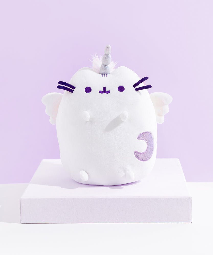 The Super Pusheenicorn Plush on top of a white square pedestal, in front of a light purple background. The plush features an all-white Pusheen sitting upright with a unicorn horn in between her ears, white plush wings, and a light purple sparkly moon on the lower right of her front body. Super Pusheenicorn has two nub paws up in the air and two nub paws at the bottom, and all embroidered details such as her eyes, mouth and head stripes, and her felt whiskers are all a dark purple.