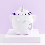 The Super Pusheenicorn Plush on top of a white square pedestal, in front of a light purple background. The plush features an all-white Pusheen sitting upright with a unicorn horn in between her ears, white plush wings, and a light purple sparkly moon on the lower right of her front body. Super Pusheenicorn has two nub paws up in the air and two nub paws at the bottom, and all embroidered details such as her eyes, mouth and head stripes, and her felt whiskers are all a dark purple.