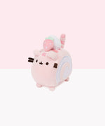 The Swiss Roll Mini Squisheen in front of white and pink background. The plush is Pusheen as a slice of a pink Swiss Roll Cake, laying on her stomach. All of Pusheen’s stripes are a dark pink, and on top of her back two stripes is a dollop of cream and a single strawberry. On the side of the plush is a white swirl filling, embroidered into the plush.