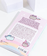 The back of the Many Lives of Pusheen the Cat book resting at an angle on top of a white background, with white cubes above the book. The back cover features copy, illustrations of Pusheenicorn, Pusheenosaurus and Mermaid Pusheen, and a rainbow at the bottom with the barcode and publisher information.