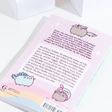 The back of the Many Lives of Pusheen the Cat book resting at an angle on top of a white background, with white cubes above the book. The back cover features copy, illustrations of Pusheenicorn, Pusheenosaurus and Mermaid Pusheen, and a rainbow at the bottom with the barcode and publisher information.