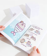 A model holding the book open in front of a white background, showing off the pages featuring Mermaid Pusheen alongside the comic ‘How To Tell If Your Cat is a Mermaid’.