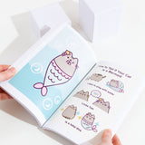 A model holding the book open in front of a white background, showing off the pages featuring Mermaid Pusheen alongside the comic ‘How To Tell If Your Cat is a Mermaid’.