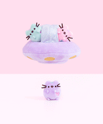 An animated gif of a plush UFO with a pastel green and pink mini Pusheen plush inside, floating over a mini pastel purple Pusheen. All of the mini plush features Pusheen sitting upright, with a pair of paws by her chest and a pair at the bottom, and an embroidered heart in the front lower right.  The bottom of the ship has embroidered yellow circles and a small ridge in the middle.