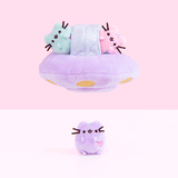 An animated gif of a plush UFO with a pastel green and pink mini Pusheen plush inside, floating over a mini pastel purple Pusheen. All of the mini plush features Pusheen sitting upright, with a pair of paws by her chest and a pair at the bottom, and an embroidered heart in the front lower right.  The bottom of the ship has embroidered yellow circles and a small ridge in the middle.