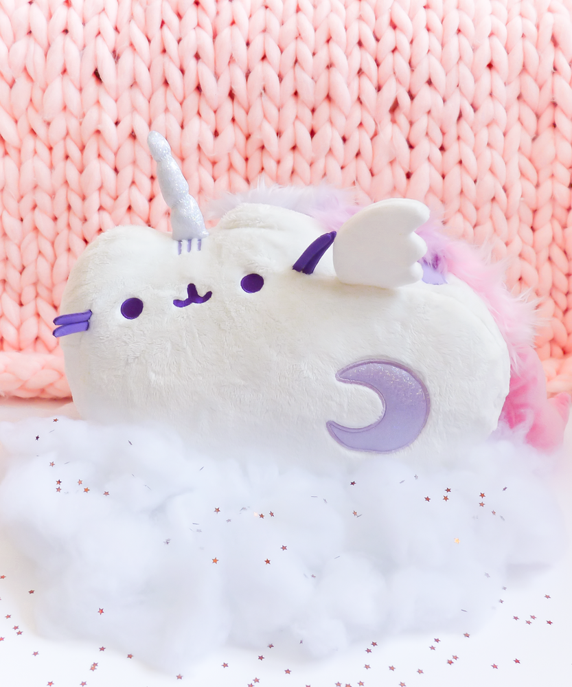 The Super Musical Pusheenicorn Plush couched in the cotton ball clouds, dotted with shiny silver star confetti. The plush is in front of a large piece of pink knit fabric.