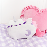 The Super Musical Pusheenicorn Plush nestled on top of a purple and white plaid blanket, pink heart pillow outlined with ruffles behind her.