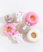 Top view of the four mini squishys, posed among three donuts with pink and white frosting. The two Hello Kitty squishys and Pusheen holding a milk jug are laid out on their backs, while Pusheen holding a donut is standing upright.