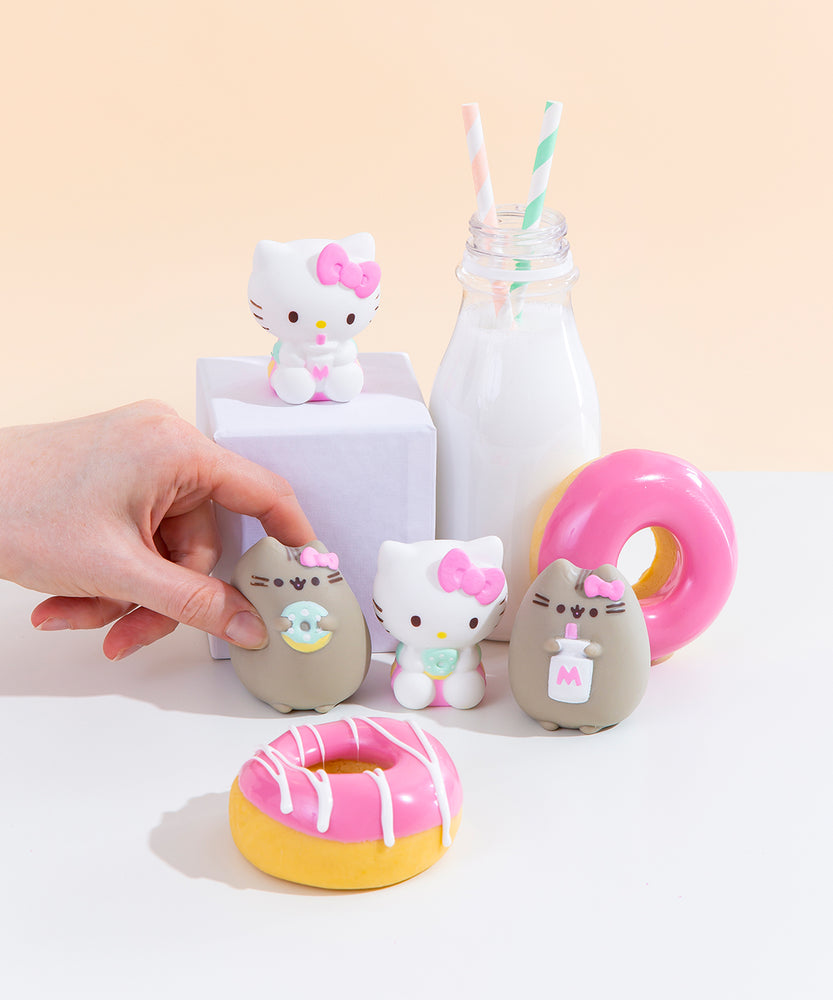 The four Squishy designs standing among pink donuts and a milk jug with two striped straws. Hello Kitty holding a milk jug is on a white podium next to the milk jug, towering above the other three designs. A hand emerging from the left has begun to squish Pusheen holding a donut in the bottom left