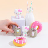 The four Squishy designs standing among pink donuts and a milk jug with two striped straws. Hello Kitty holding a milk jug is on a white podium next to the milk jug, towering above the other three designs. A hand emerging from the left has their fingers around Pusheen holding a donut in the bottom left, preparing to squish.
