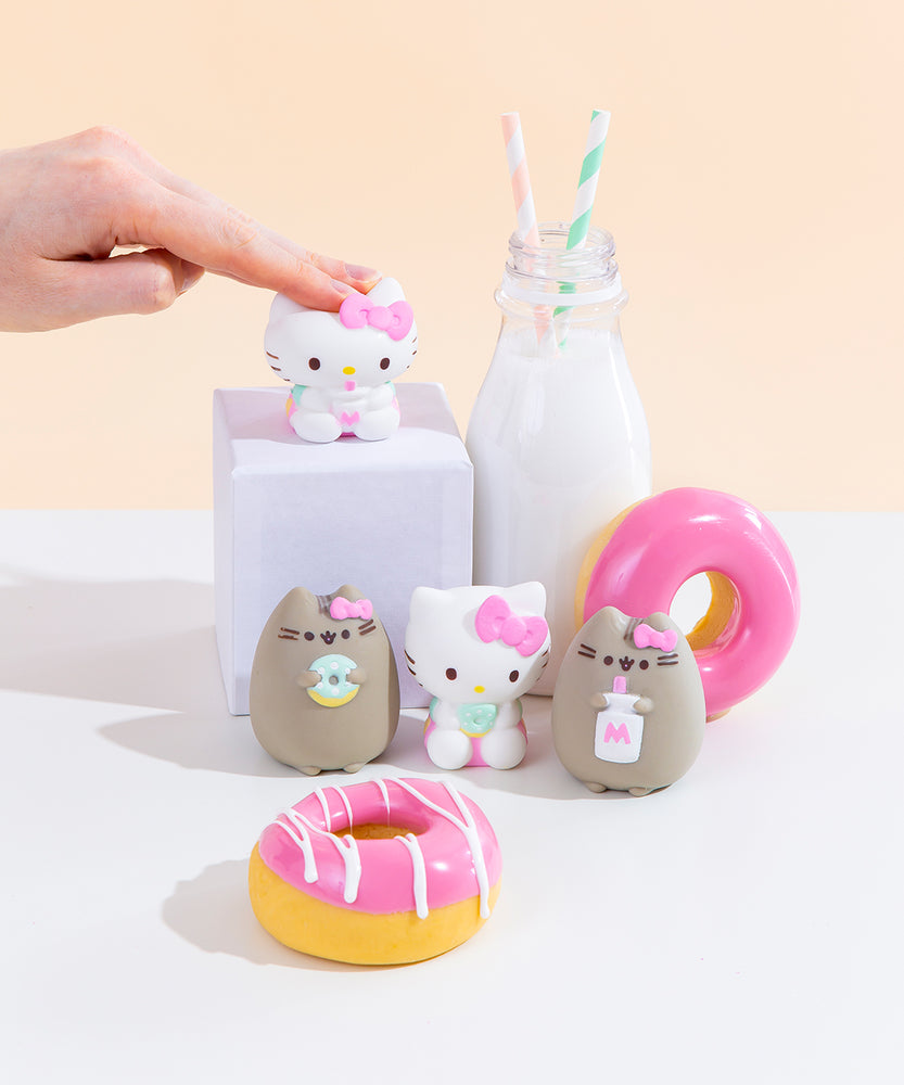 The four Squishy designs standing among pink donuts and a milk jug with two striped straws. Hello Kitty holding a milk jug is on a white podium next to the milk jug, towering above the other three designs. A hand emerging from the left is patting the Hello Kitty on the podium hard enough to squish her.