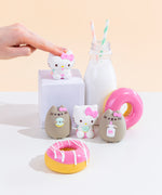 The four Squishy designs standing among pink donuts and a milk jug with two striped straws. Hello Kitty holding a milk jug is on a white podium next to the milk jug, towering above the other three designs. A hand emerging from the left is patting the Hello Kitty on the podium hard enough to squish her.