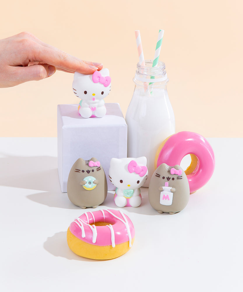 The four Squishy designs standing among pink donuts and a milk jug with two striped straws. Hello Kitty holding a milk jug is on a white podium next to the milk jug, towering above the other three designs. A hand emerging from the left is patting the Hello Kitty on the podium with their fingers.