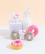 The four Squishy designs standing among pink donuts and a milk jug with two striped straws. The four possible surprise squishy designs include 2 designs of Hello Kitty sitting and 2 designs of Pusheen wearing Hello Kitty’s pink bow, with them either holding a milk jug with a straw or a mint donut.