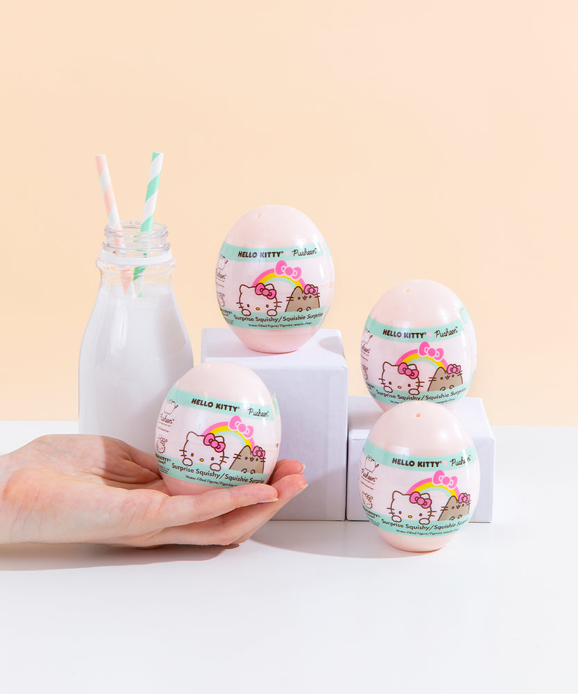Four light pink plastic egg containers for the surprise squishies, with a white and mint wrapping featuring the Hello Kitty x Pusheen logo, posed on top of various white podiums next to a milk jug with two striped straws. A hand coming from the left is holding the bottom left egg container, which fits in the palm of their hand.