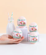 Four light pink plastic egg containers for the surprise squishies, with a white and mint wrapping featuring the Hello Kitty x Pusheen logo, posed on top of various white podiums next to a milk jug with two striped straws. A hand coming from the left is holding the bottom left egg container, which fits in the palm of their hand.