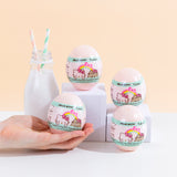 Four light pink plastic egg containers for the surprise squishies, with a white and mint wrapping featuring the Hello Kitty x Pusheen logo, posed on top of various white podiums next to a milk jug with two striped straws. A hand coming from the left is holding the bottom left egg container, which fits in the palm of their hand.