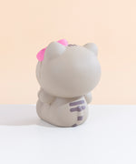 Back view of the Hello Kitty Jumbo Squishy facing to the left, seated on a white floor in front of a pale yellow background. The back of Hello Kitty’s Pusheen costume has two dark grey stripes on the back, along with a striped tail sculpted into the squishy that points upwards her back.