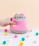 Front view of the Pusheen Jumbo Squishy seated among colorful puff balls in front of a pale yellow background. A model’s hand is squeezing in on the squishy from the left.