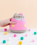 Front view of the Pusheen Jumbo Squishy seated among colorful puff balls in front of a pale yellow background, a model’s hand holding onto the squishy from the left. The Pusheen Jumbo Squishy is wearing a pink dress with yellow pockets, mint sleeves and a collar, as well as Hello Kitty's bow on top of her head. Pusheen’s left paw is raised up close to her face.