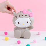 Front view of the Hello Kitty Jumbo Squishy seated among colorful puff balls in front of a pink background. A model’s hand is strongly pinching the Jumbo Squishy’s left ear.