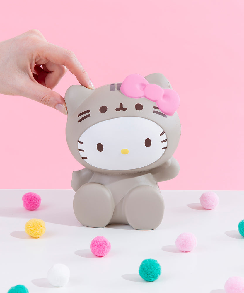 Front view of the Hello Kitty Jumbo Squishy seated among colorful puff balls in front of a pink background, a model’s hand lightly pinching her left ear. Hello Kitty is seated with her legs on the ground, waving her right arm. She is wearing a grey Pusheen costume that covers her entire body, and the hood of the costume features Pusheen's face and whiskers. A plush pink bow sits on top of the right ear.