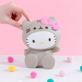 Front view of the Hello Kitty Jumbo Squishy seated among colorful puff balls in front of a pink background, a model’s hand lightly pinching her left ear. Hello Kitty is seated with her legs on the ground, waving her right arm. She is wearing a grey Pusheen costume that covers her entire body, and the hood of the costume features Pusheen's face and whiskers. A plush pink bow sits on top of the right ear.