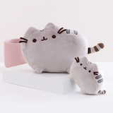 The Medium Pusheen Plush on a white square platform, with a mini Pusheen plush angled in front as a size comparison for the Medium Plush. The two plush are in front of a pink ribbed pot and a light grey background.