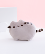 The Medium Pusheen Plush on top of a white floor, in front of a pink background and two white shapes, a big square and a small hexagon. The Pusheen Plush features a grey cat with a few dark stripes standing in profile. The front and back left legs are visible from the front, both ears, her felt whiskers, and her embroidered face are faced directly to the front, and a small striped tail sticks out immediately from the side. The plush is very rounded out, with no hard edges.