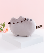 The Medium Pusheen plush on top of a white square platform. A pink peony is visible on the left in the background, and the plush is in front of a light pink background.