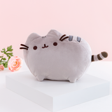 The Medium Pusheen plush on top of a white square platform. A pink peony is visible on the left in the background, and the plush is in front of a light pink background.