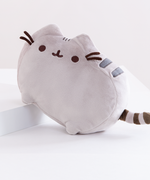 The Pusheen Plush angled upwards by having her front legs rest on top of a white block. The plush has three small dark grey stripes embroidered in-between her ears, and two thick dark grey stripes embroidered on her side, behind her right ear and right felt whiskers. Her tail features 3 dark brown stripes.