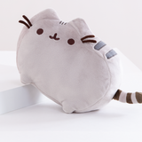 The Pusheen Plush angled upwards by having her front legs rest on top of a white block. The plush has three small dark grey stripes embroidered in-between her ears, and two thick dark grey stripes embroidered on her side, behind her right ear and right felt whiskers. Her tail features 3 dark brown stripes.