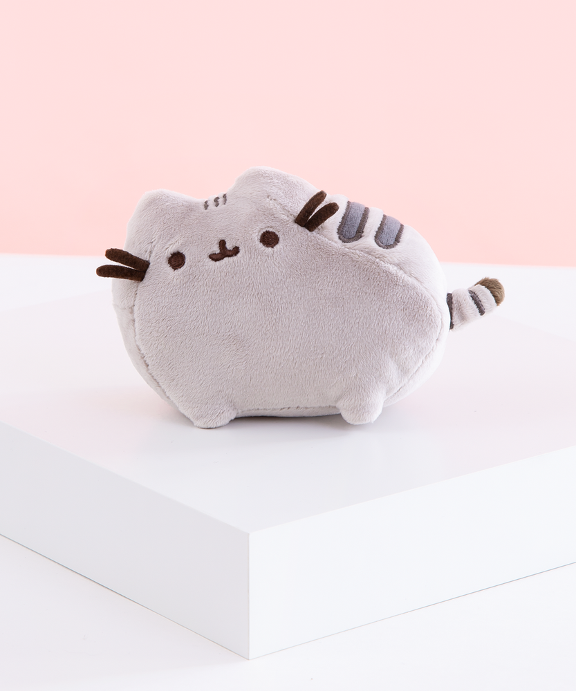 The Medium Pusheen plush on top of a white square platform, in front of a white and pink background.
