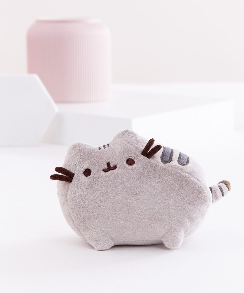 The Mini Pusheen Plush on top of a white floor, in front of a white background with a pink vase and two white shapes, a big square and a small hexagon. The mini Pusheen Plush features a grey cat with a few dark stripes standing in profile. The front and back left legs are visible from the front, both ears, her felt whiskers, and her embroidered face are faced directly to the front, and a small striped tail sticks out immediately from the side.