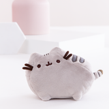 The Mini Pusheen Plush on top of a white floor, in front of a white background with a pink vase and two white shapes, a big square and a small hexagon. The mini Pusheen Plush features a grey cat with a few dark stripes standing in profile. The front and back left legs are visible from the front, both ears, her felt whiskers, and her embroidered face are faced directly to the front, and a small striped tail sticks out immediately from the side.