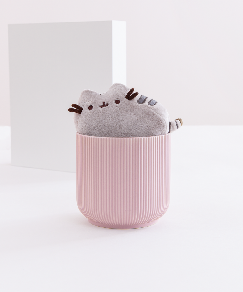 The Mini Pusheen Plush inside a round pink ribbed pot, in front of a white background with a square in the background. The mini plush fits snugly in the rim of the small pot.