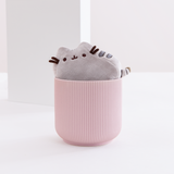 The Mini Pusheen Plush inside a round pink ribbed pot, in front of a white background with a square in the background. The mini plush fits snugly in the rim of the small pot.