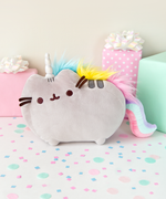 The Pusheenicorn plush  on top of a white floor covered in confetti, surrounded by gifts. The Pusheenicorn Plush stands in profile similar to the standard plush, this time with a plush sparkly white unicorn horn inbetween her ears, tufts of blue, yellow and pink fur along her back as a mane, and a long, pink/mint/lilac plush tail that curves down to the floor.