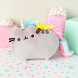 The Pusheenicorn plush  on top of a white floor covered in confetti, surrounded by gifts. The Pusheenicorn Plush stands in profile similar to the standard plush, this time with a plush sparkly white unicorn horn inbetween her ears, tufts of blue, yellow and pink fur along her back as a mane, and a long, pink/mint/lilac plush tail that curves down to the floor.