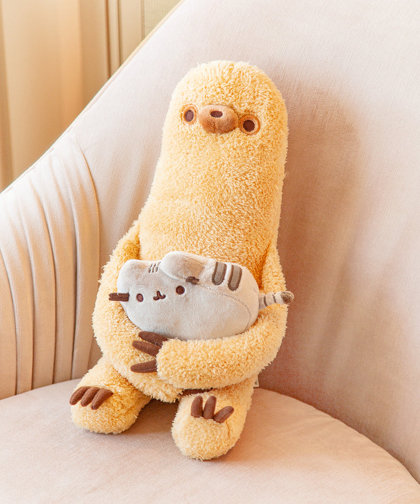 Sloth holding a mini Pusheen plush, sitting in a big beige armchair. Sloth is a golden brown rounded rectangle with long arms and legs that have three brown claws each. Sloth’s arms are long enough to hold a mini Pusheen in his lap. Sloth’s fur is lightly fuzzy, and his face features a round snoot with a brown dot nose, and two dot eyes with light brown rings around them. 