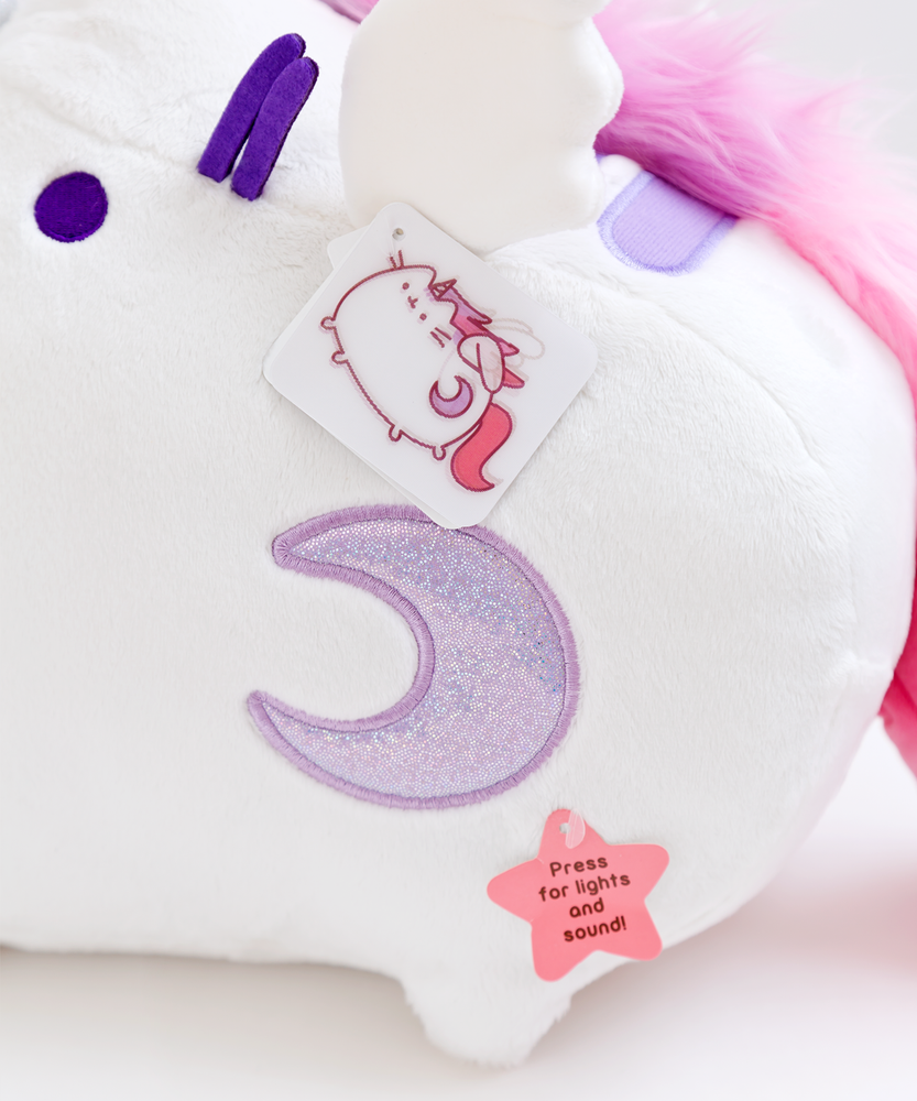 Close up of the light purple sparkly crescent moon embroidered into the plush. There is a lenticular hangtag hanging from the wing featuring Super Pusheenicorn flapping her wings. Underneath the moon is a paper star hangtag reading ‘Press for lights and sound!’