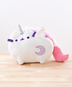 The plush features an all-white Pusheen standing on her legs, a unicorn horn in between her ears. She has white plush wings around the edge of her top back stripe, and a light purple sparkly moon on the lower right of her front body. Super Pusheenicorn’s embroidered details such as her eyes, mouth and head stripes, and her felt whiskers are all a dark purple. She has a fluffy multicolor light purple, purple and pink mane running down her back, leading to her solid pink plush tail that curves upwards